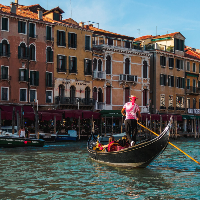 Una coppia è seduta in gondola, condotta a remi da un gondoliere vestito con il tipico cappello e la maglia a righe bianche e rosse orizzontali.
